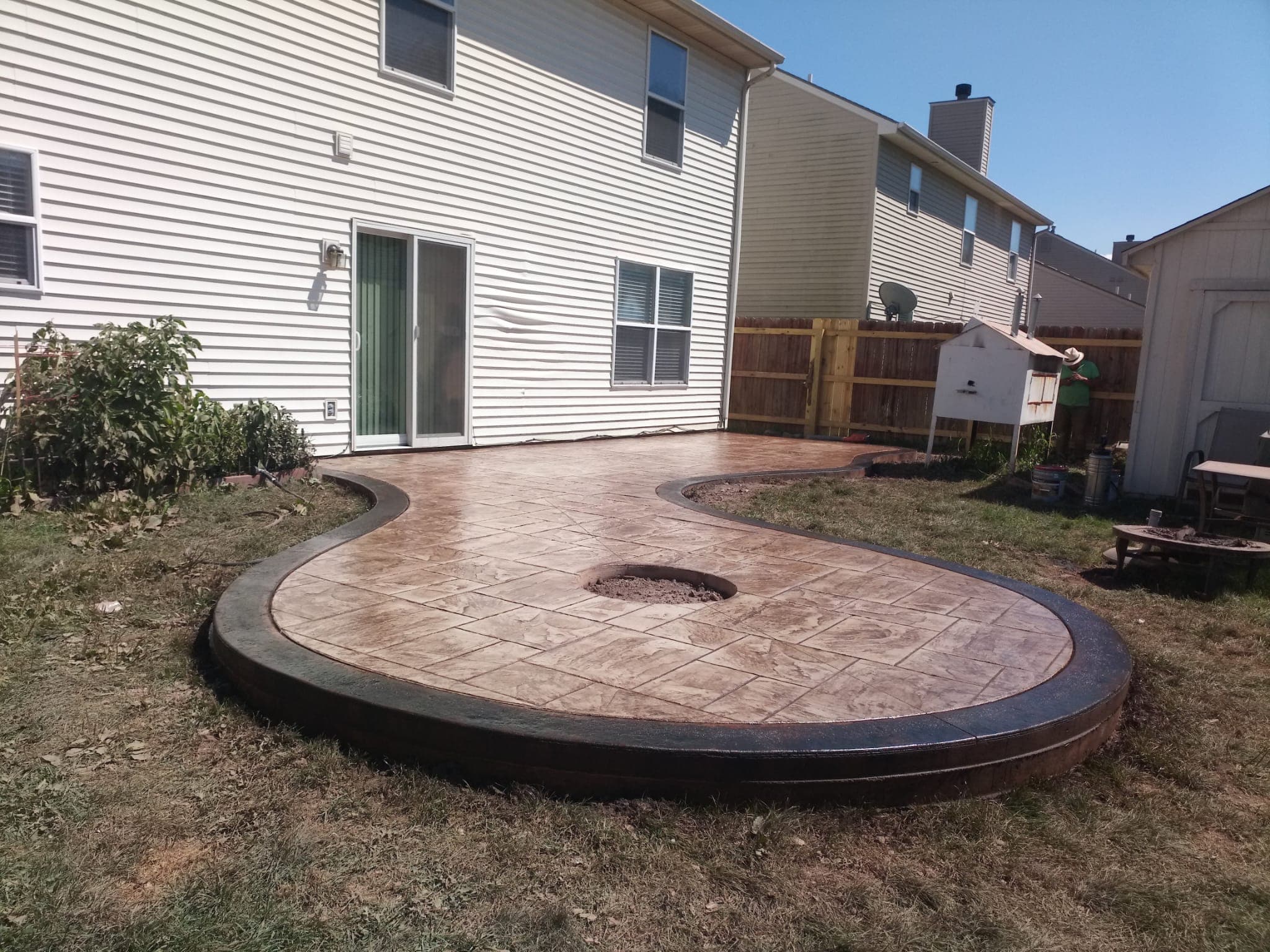 Completed stamped concrete patio with a curved outline, dark border, and circular opening, extending into the backyard.