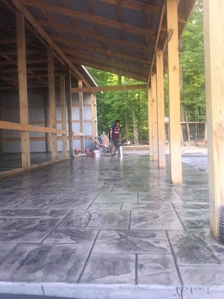 Gray concrete with rectangular stone-like stamp impressions beneath an open timber-framed structure, with a worker in the distance.